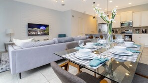 Una sala de estar moderna con una mesa de vidrio dispuesta para cuatro comensales, un sofá gris y una cocina al fondo.