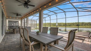 Une terrasse extérieure avec une table et des chaises, un ventilateur de plafond, des guirlandes lumineuses et une vue sur un lac et une forêt.