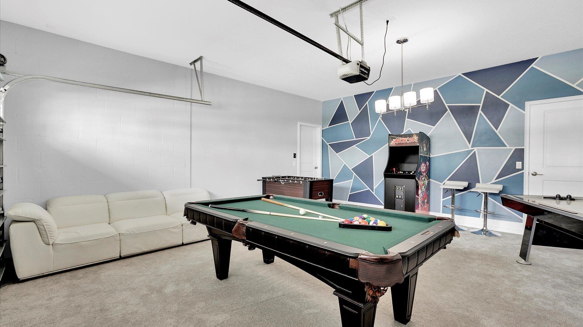 A pool table with a green felt surface, a white sofa, and a foosball table in a room with a geometric-patterned wall and a projector mounted on the ceiling.
