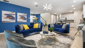 Ein modernes Wohnzimmer mit einem blauen Sofa, einem grauen Sessel und einem runden Couchtisch. Eine Treppe führt zu einer Küchenzeile mit weißen Schränken.