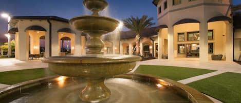 A fountain in a courtyard with a building featuring arched walkways and a pool area.