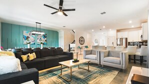 A modern living room with a black sofa, grey armchairs, and a coffee table. There is a dining area with a table set for six. The kitchen is visible in the background with white cabinets and stainless steel appliances. - Last Minute Disney! Private Pool, Hot Tub, Game Room & Themed Bedrooms. Book Now! #87159 (Orlando)