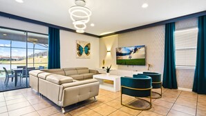 Uma sala de estar moderna com um sofá seccional, uma mesa de centro e duas poltronas de destaque. Há uma TV de tela plana na parede, uma planta decorativa e uma janela com cortinas.