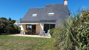 Een huis met een schuin dak, een overdekte patio met een tafel en stoelen, en een tuin.