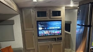 Ein kompakter Wohnbereich mit einem an einer Holzkommode befestigten Flachbildfernseher, einem Sofa mit einem orangefarbenen Kissen, einem gemusterten Teppich und einem Kühlschrank.