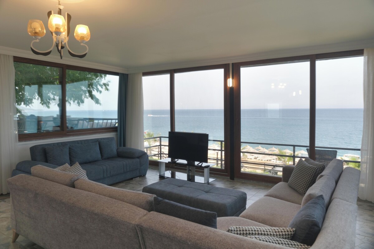 Een ruime woonkamer met een grote bank, een televisie en een balkon met uitzicht op zee.