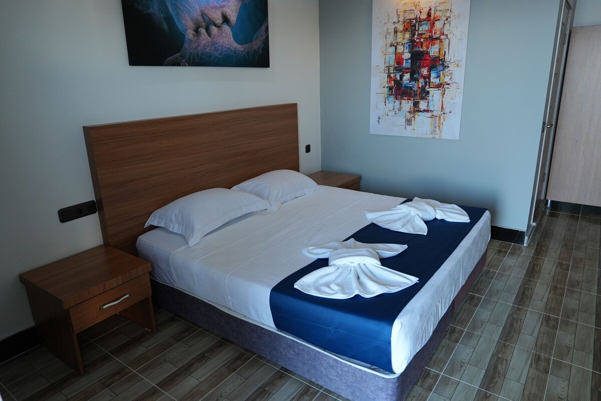Een net opgemaakt bed met witte lakens en een blauw accent, een houten nachtkastje met een lade, en een groot abstract schilderij aan de muur.