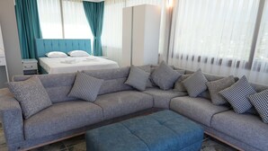 Moderni hotellihuone, jossa on harmaa kulmasohva, sininen jalkarah, valkoisilla lakanoiden peitetty sänky, turkoosit verhot ja suuri ikkuna, jossa on läpikuultavat valkoiset verhot.