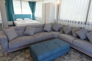 Een moderne hotelkamer met een grijze modulaire bank, een blauwe poef, een bed met witte lakens, turquoise gordijnen en een groot raam met dunne witte gordijnen.