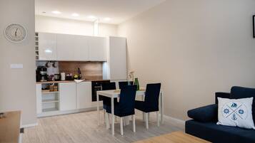 Una sala moderna con un sofá azul oscuro y una mesa de centro de madera, un comedor con una mesa y sillas blancas, y una cocina con gabinetes blancos y un horno empotrado.