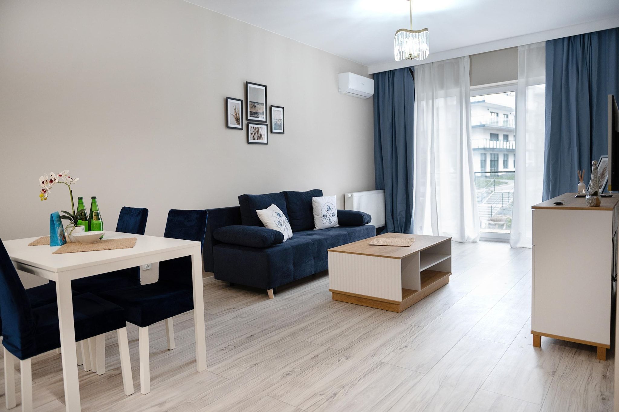 Una sala de estar moderna con un sofá, una mesa de comedor y una mesa de centro. Hay cuadros enmarcados en la pared, una ventana con cortinas y un equipo de aire acondicionado.