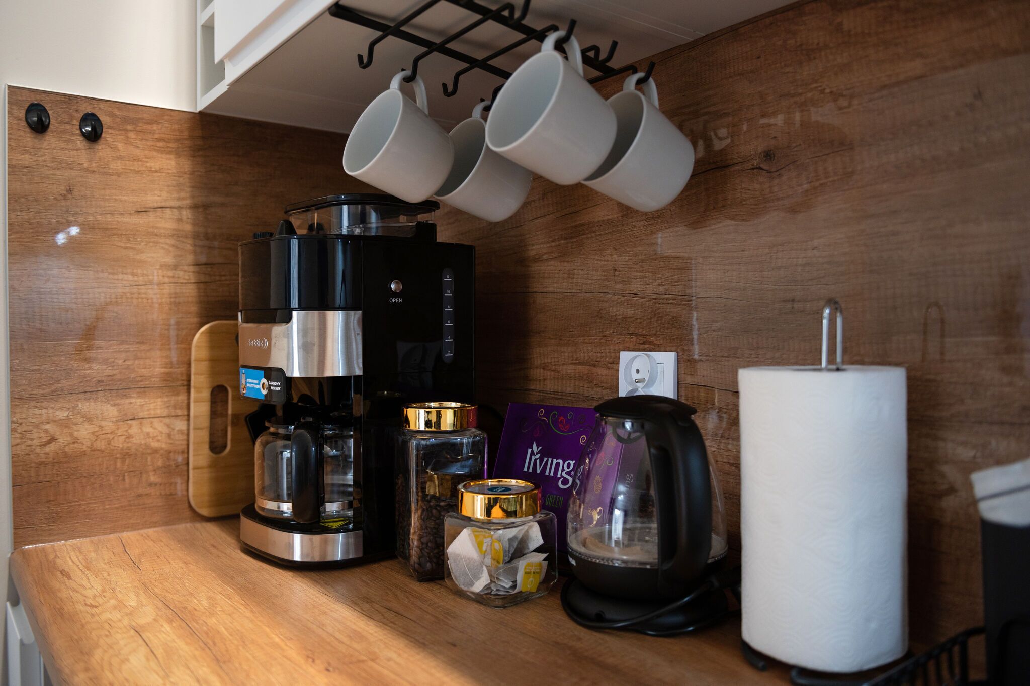 Un piano di lavoro in cucina con una macchina per il caffè, tazze, una scatola di tè, un bollitore e un rotolo di carta assorbente.