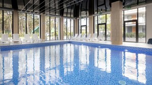 Ein Hallenbad mit klarem, blauem Wasser, umgeben von weißen Liegestühlen und großen Fenstern mit Blick ins Freie.