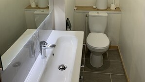 Un baño pequeño y moderno con un lavabo, inodoro y espejo blancos. Hay dos cuadros colgados en la pared que representan escenas de playa.