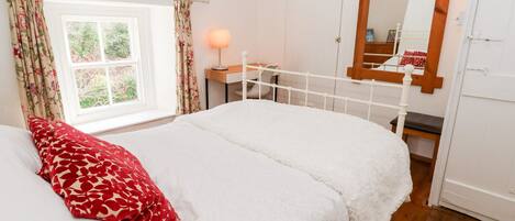 A bedroom with a bed, a nightstand, a lamp, a window with curtains, a wooden headboard, and a mirror on the wall.