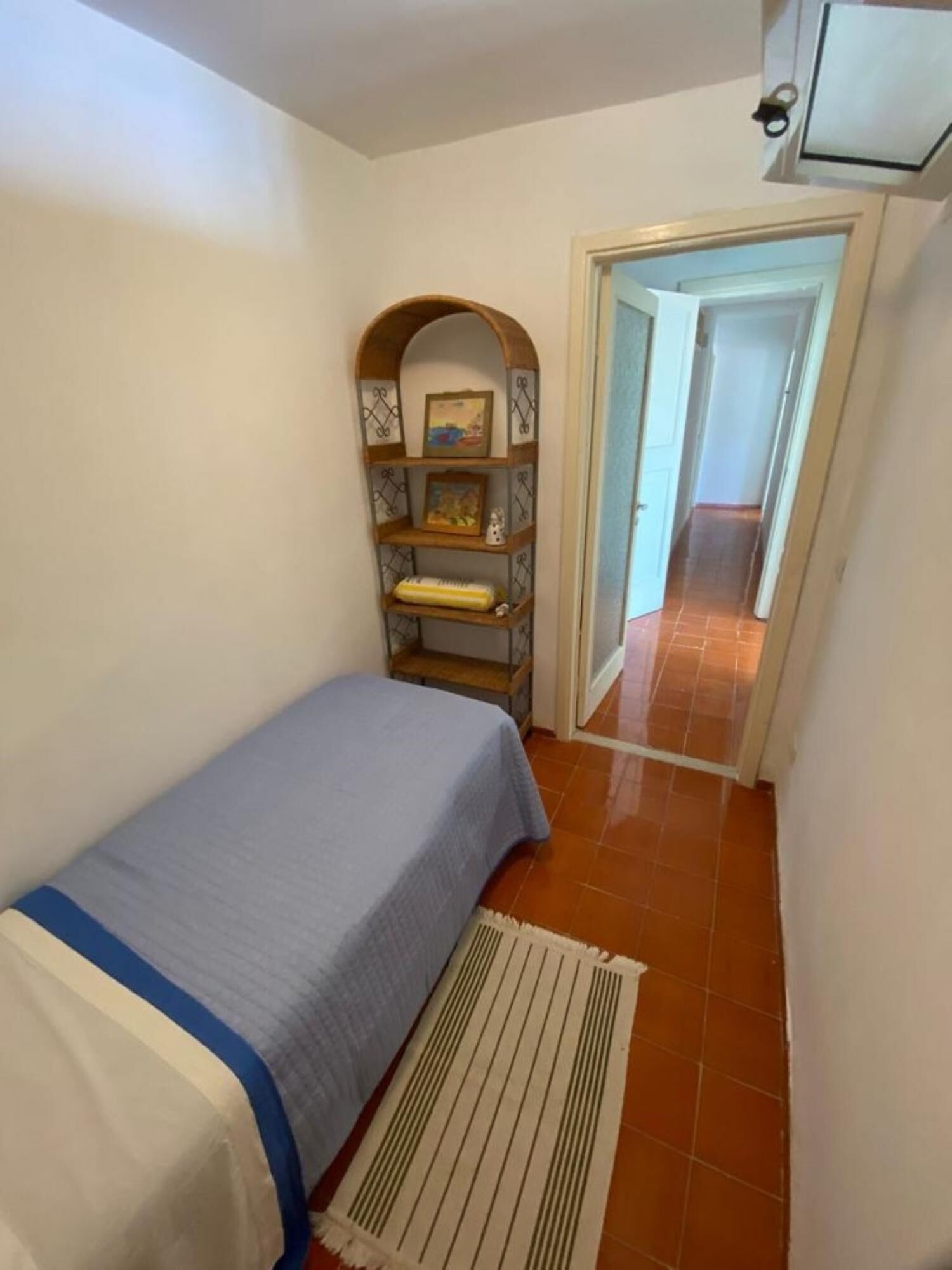 A small, narrow room with a single bed, a shelf with framed pictures, and a door leading to another room.