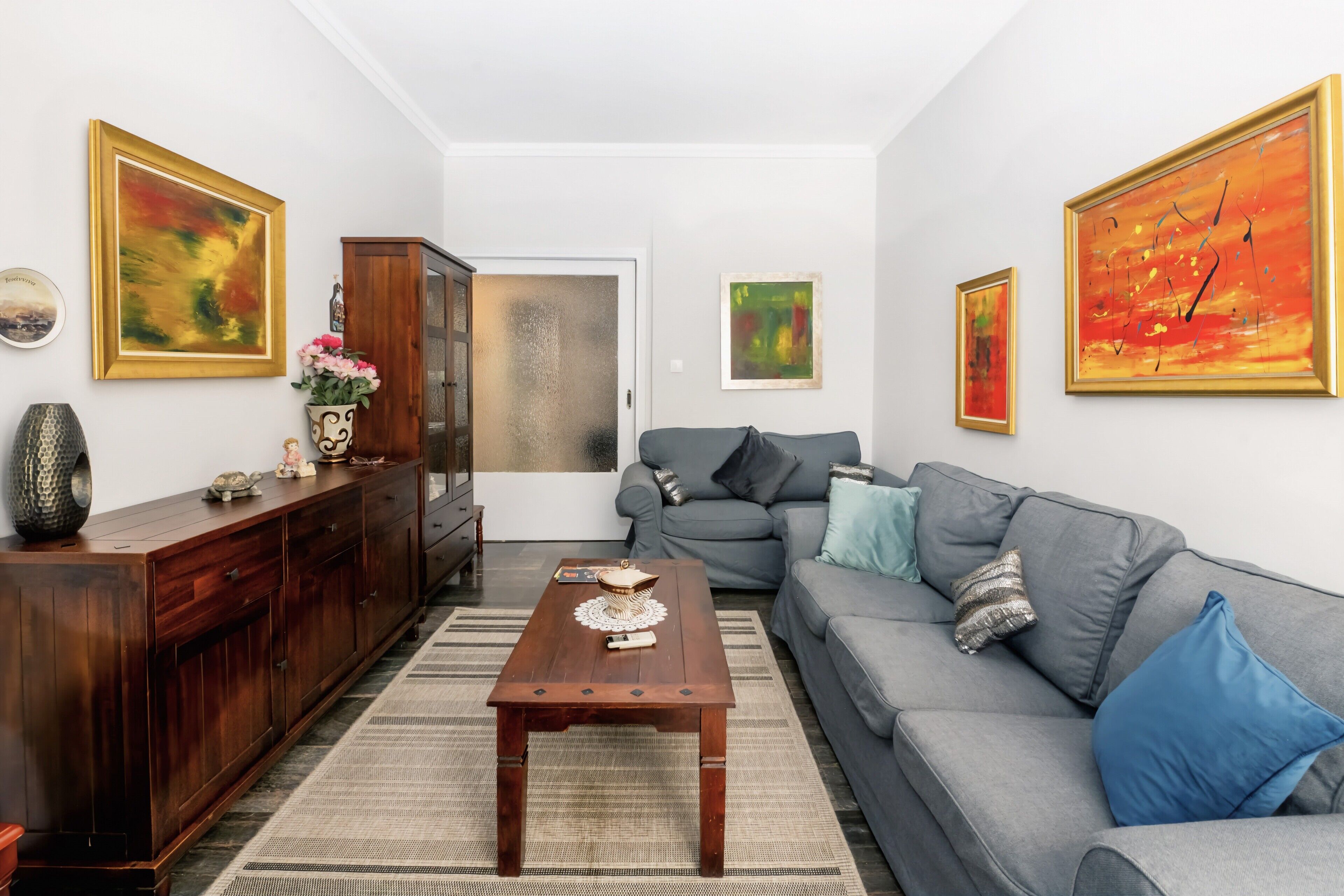 A living room with a wooden coffee table, grey sofa, and a cabinet. There are three paintings on the walls.