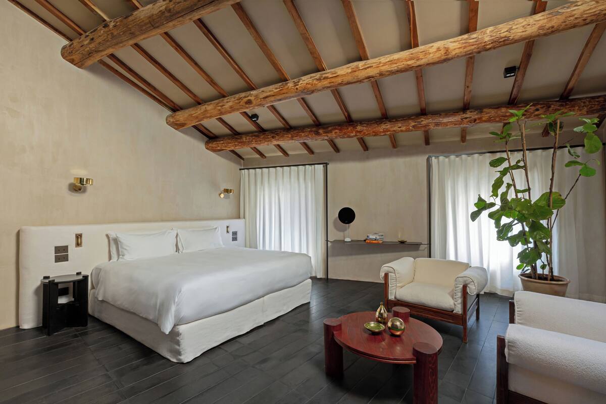 A hotel room with a large bed, a wooden ceiling, a desk with a chair, a small table with decorative items, and a potted plant.