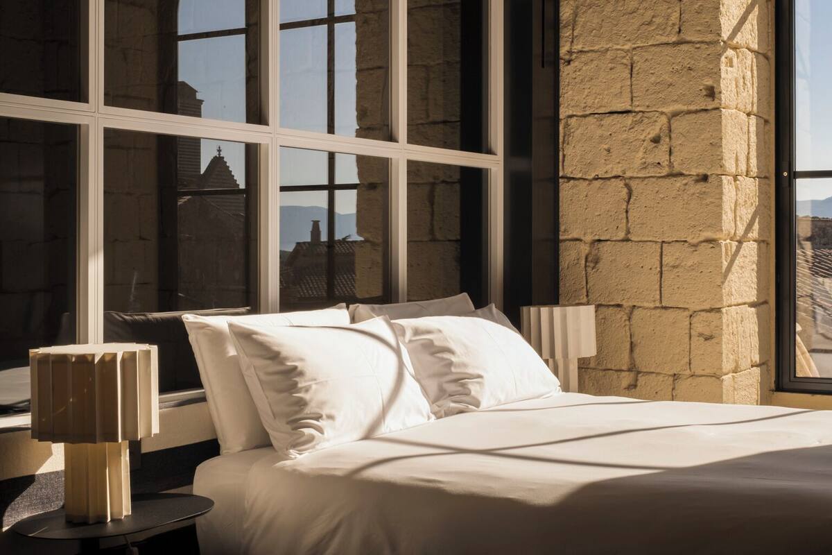 A bedroom with a large bed, a nightstand, and a lamp. There are large windows with a view of the outside.