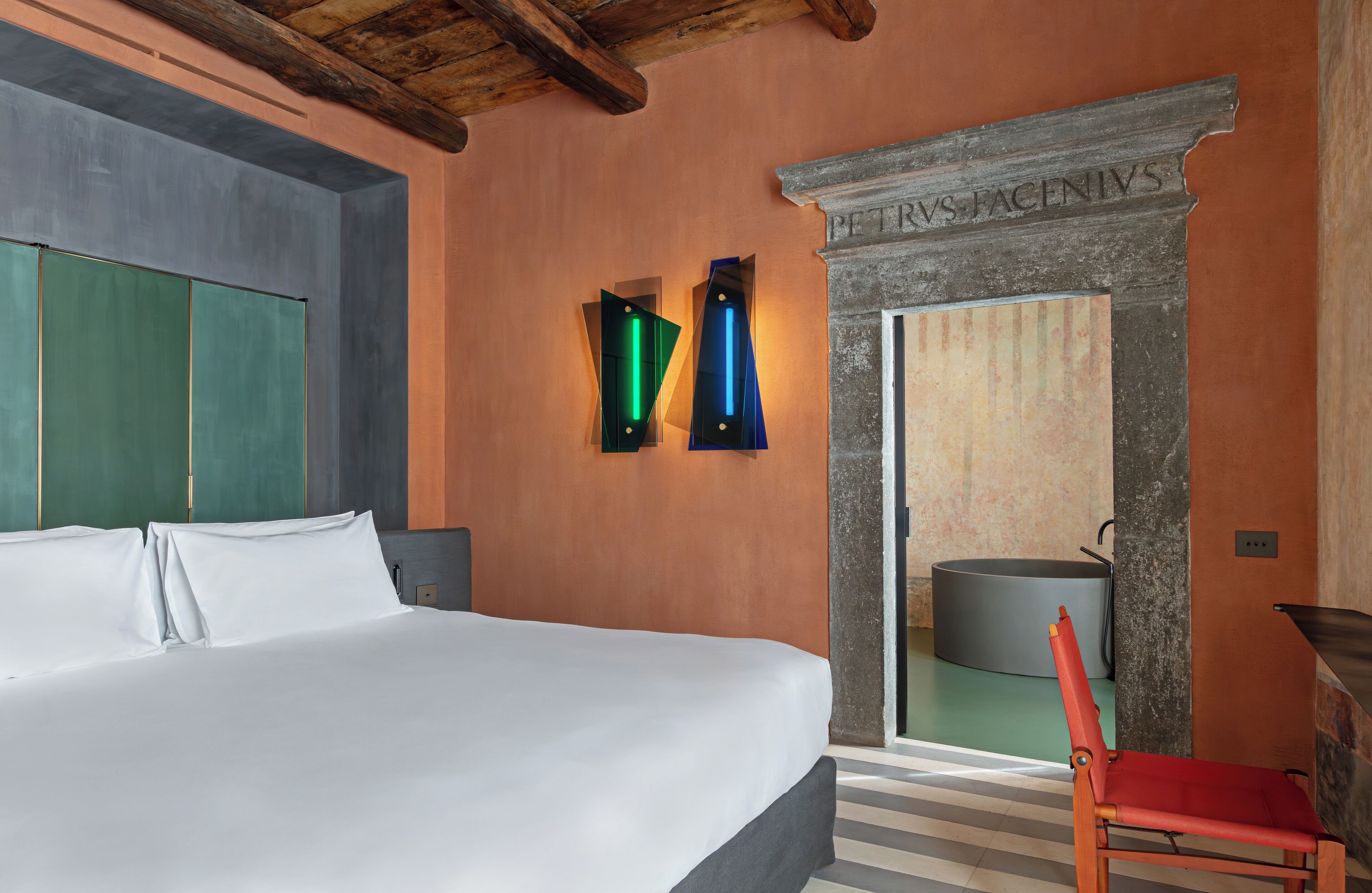Une chambre d’hôtel avec un grand lit, un fauteuil rouge et une cheminée en pierre portant l’inscription « Petrus Facenius ».