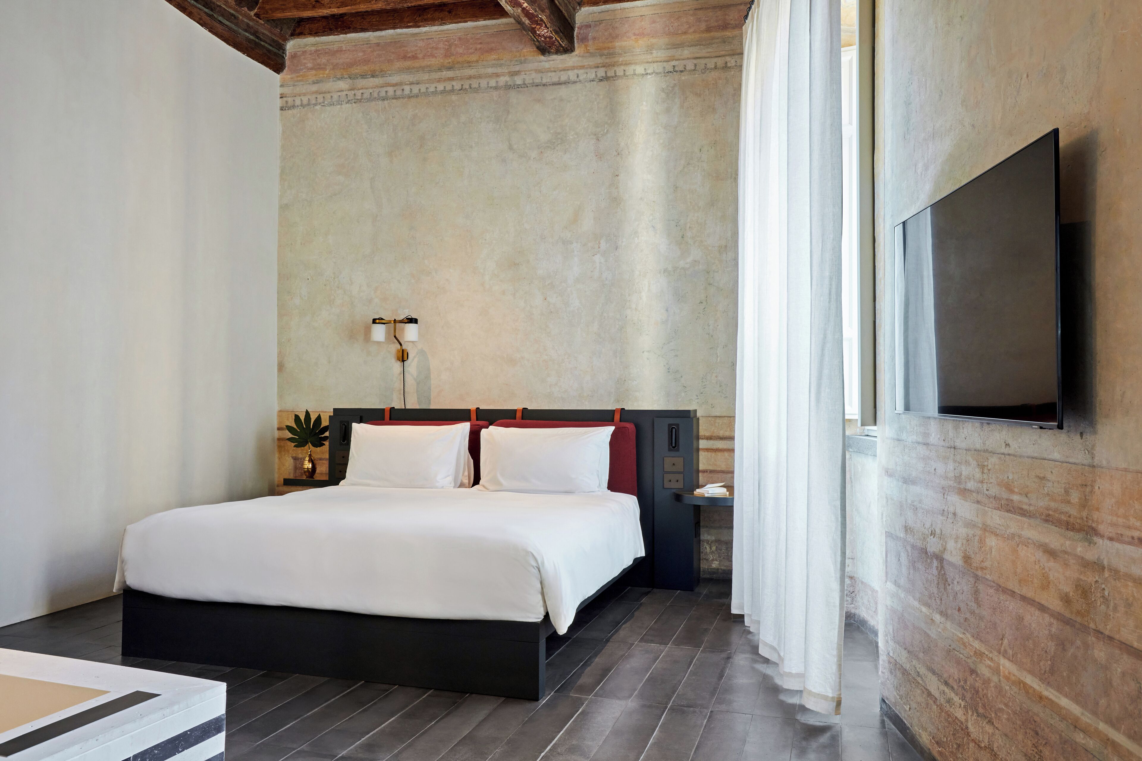 A modern hotel room with a large bed, a flat-screen TV mounted on the wall, and a small bedside table with a lamp.