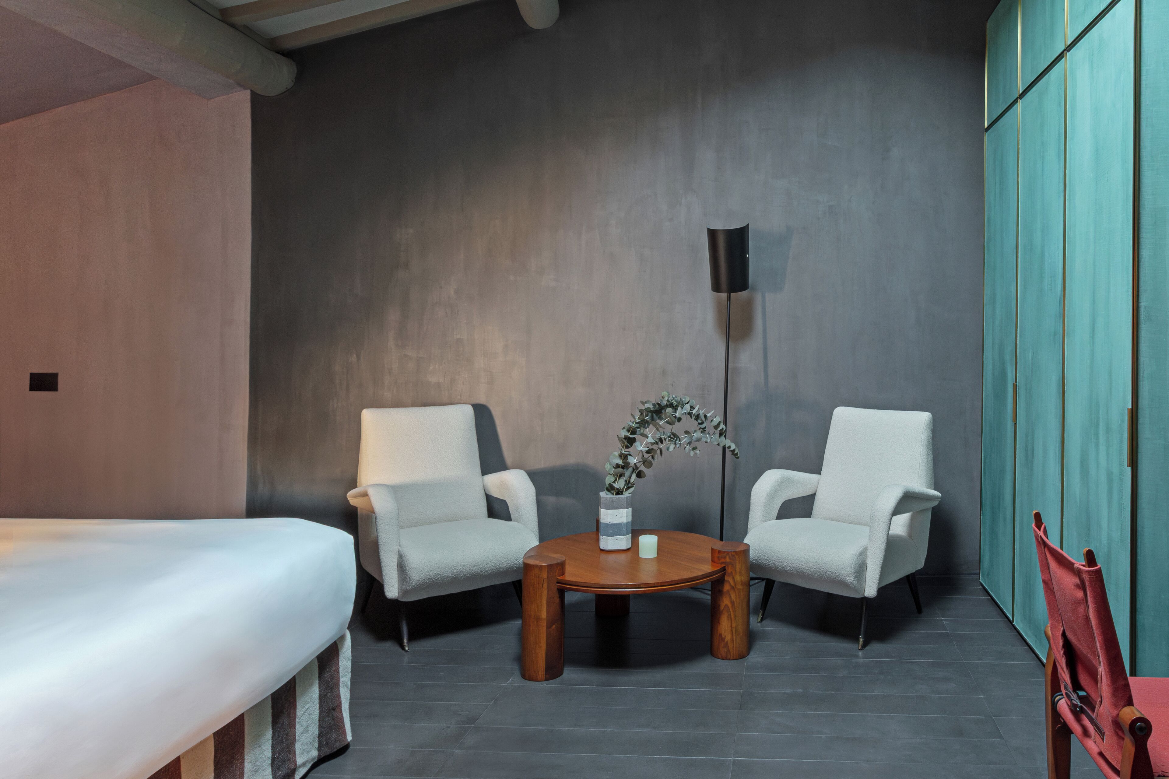 A modern room with a bed, two white armchairs, a wooden coffee table with a vase, and a tall floor lamp.