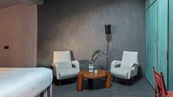 A modern room with a bed, two white armchairs, a wooden coffee table with a vase, and a tall floor lamp.