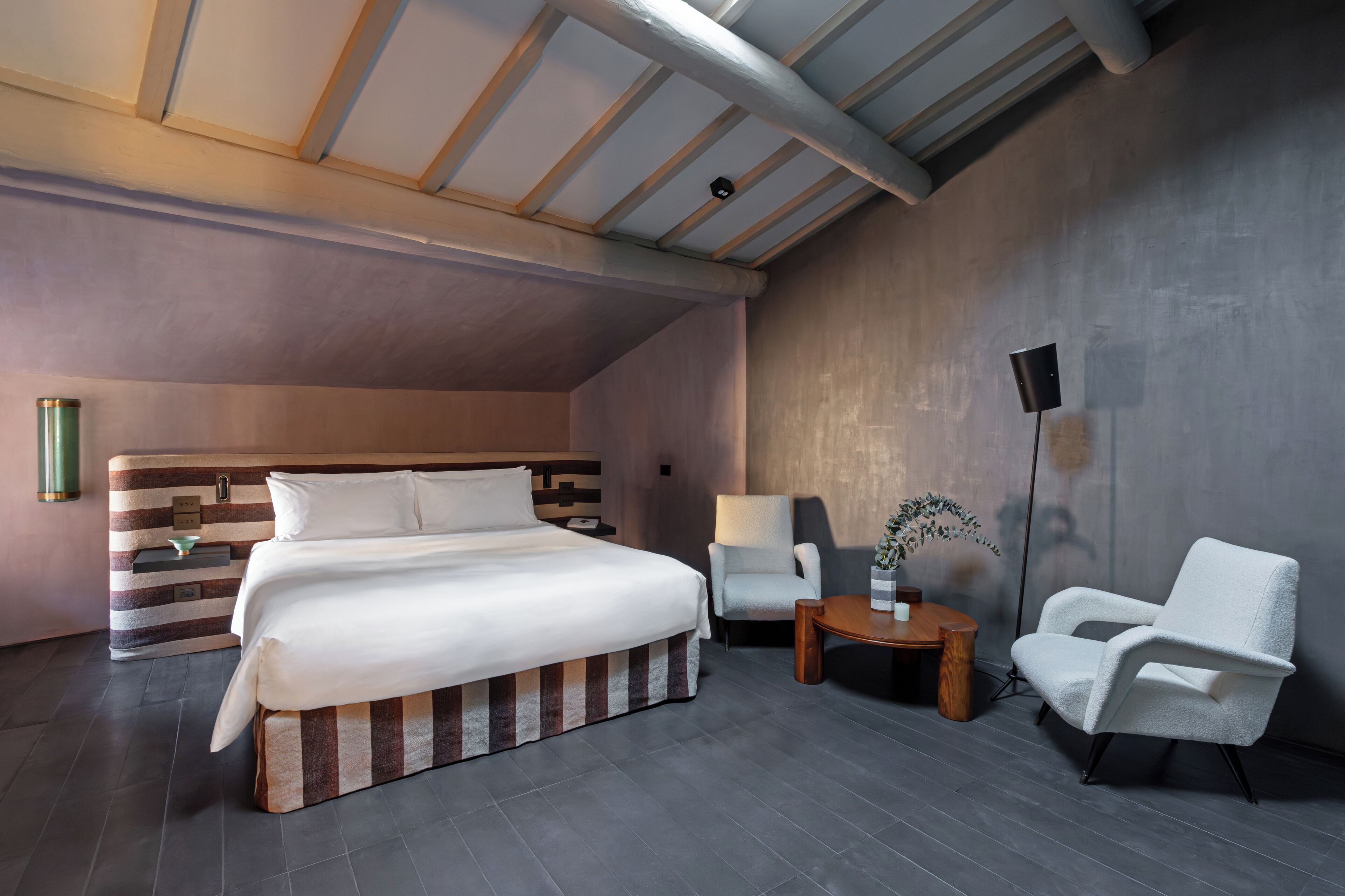 Une chambre d’hôtel moderne avec un grand lit, deux fauteuils blancs, une petite table ronde et une lampe sur pied.