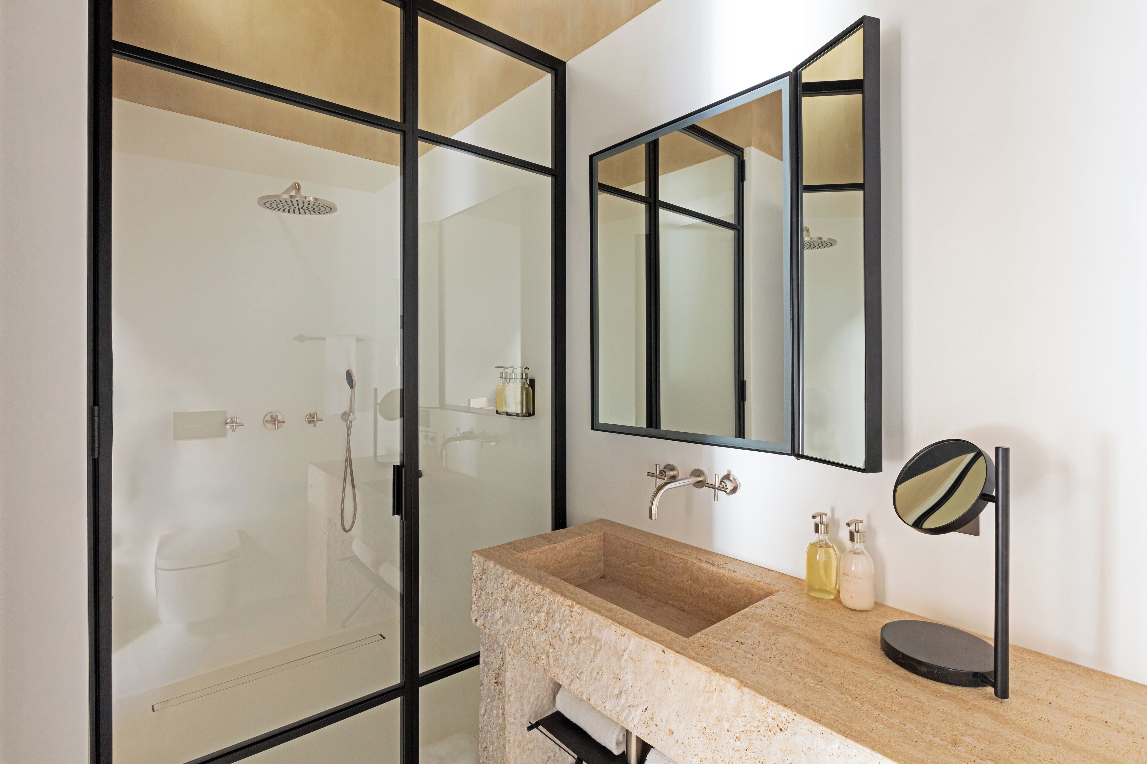 A bathroom with a glass-enclosed shower, a beige sink with a black countertop, and a mirror with a round frame.