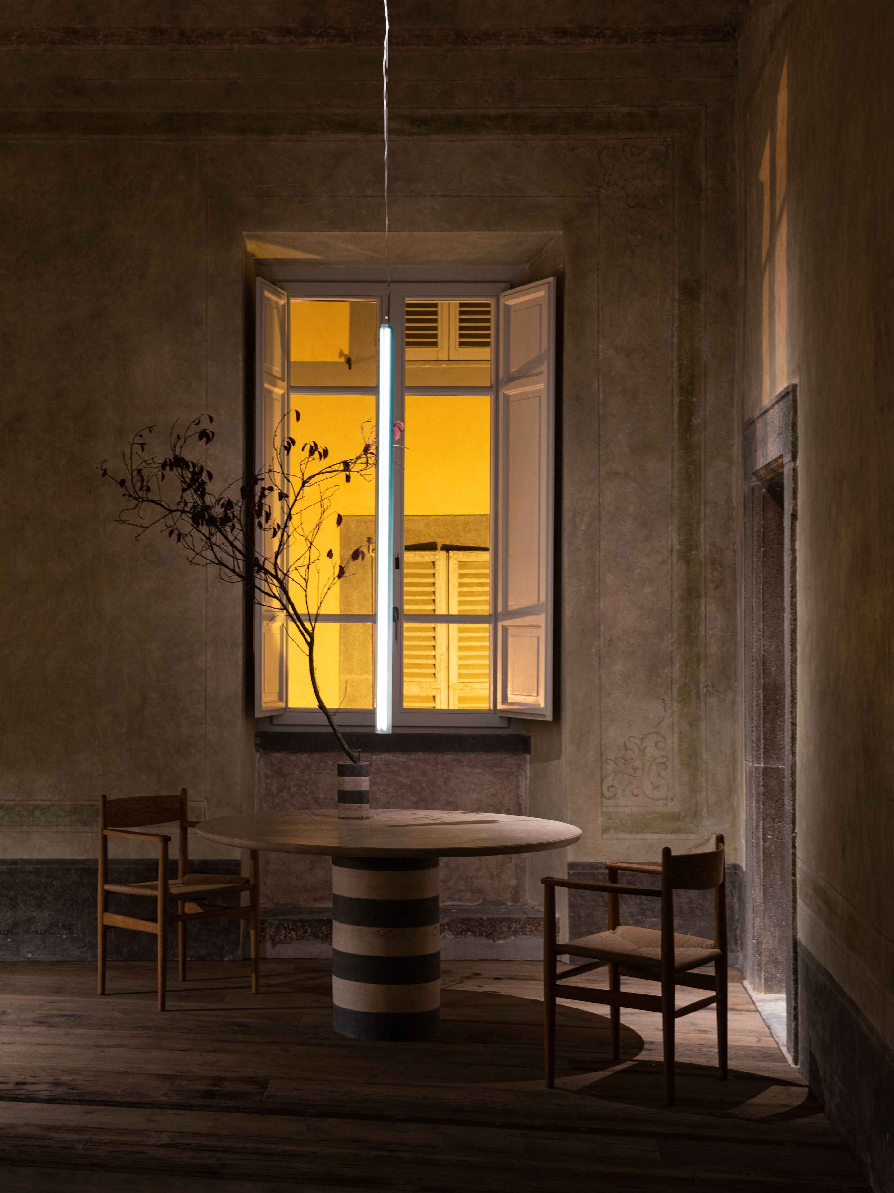 Una habitación con una mesa redonda, dos sillas, un jarrón con ramas y una ventana con contraventanas abiertas.