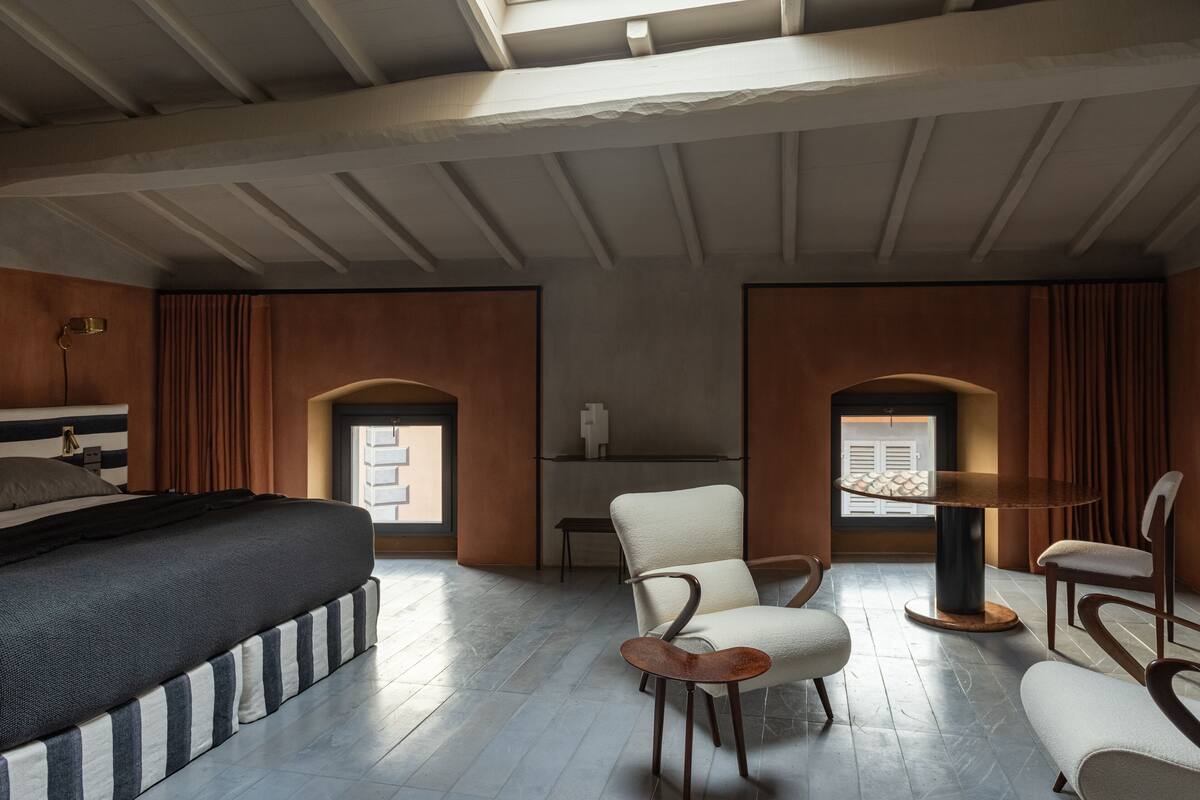 A modern bedroom with a large bed, two armchairs, a small table, and a window with shutters.