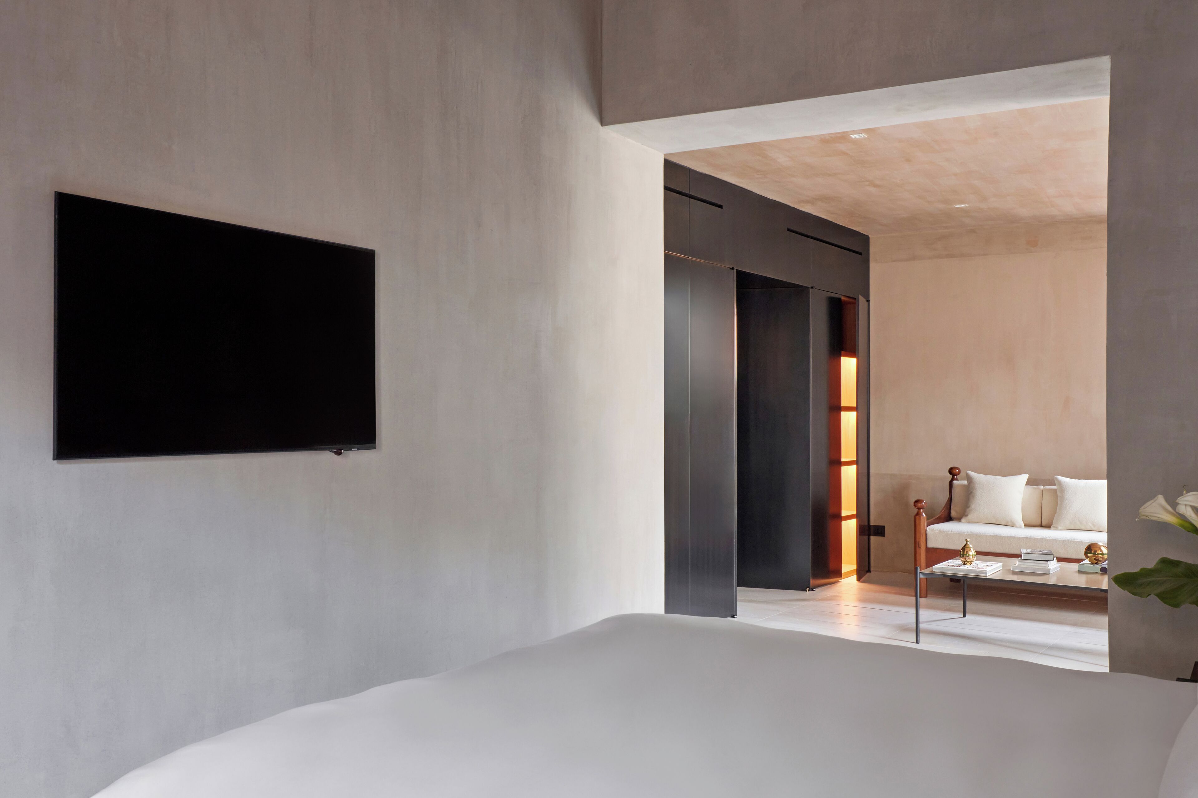Un dormitorio moderno con una cama grande, un televisor de pantalla plana en la pared y un banco de madera con cojines al fondo.