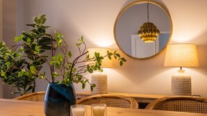 A dining area with a wooden table, two chairs, a vase with green foliage, two candles, a mirror, and two lamps.