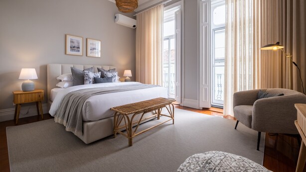 A modern bedroom with a large bed, bedside lamps, a wicker bench, a chair, and a view of the outdoors through large windows.