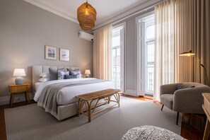 Une chambre moderne avec un grand lit, des lampes de chevet, un banc en osier, un fauteuil et une vue sur l’extérieur grâce à de grandes fenêtres.