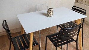 Une table à la surface blanche, entourée de quatre chaises noires aux pieds en bois. Un chapeau de paille tressé est accroché au mur, au-dessus de la table.