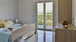 Ein Schlafzimmer mit Bett, Nachttisch, Kommode und einem Balkon mit Blick auf Grünflächen und Hügel.
