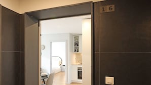 Ein modernes Interieur mit einer dunkel gefliesten Wand, einer Tür mit der Aufschrift '16' und einem sichtbaren Küchenbereich mit Holzboden.