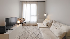 Ein modernes Wohnzimmer mit einem Flachbildfernseher auf einem weißen Schrank, einem beigefarbenen Sofa, einem gemusterten Teppich und einem Sessel mit Lampe.