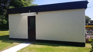 Una pequeña construcción de una sola habitación, con puerta y ventana, rodeada de césped y árboles.