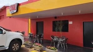 Un hôtel à la façade rouge et jaune, avec un espace extérieur aménagé avec des tables et des chaises, et un véhicule blanc garé à proximité.