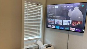 A bedroom with a bed, a TV mounted on the wall displaying a streaming service interface, a window with blinds, and a white air purifier. - New! Renovated 2BD Home in Downtown (Billings)