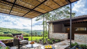 Eine Terrasse im Freien mit Korbmöbeln, einem Steintisch mit Früchten und Getränken und Blick auf grüne Hügel.