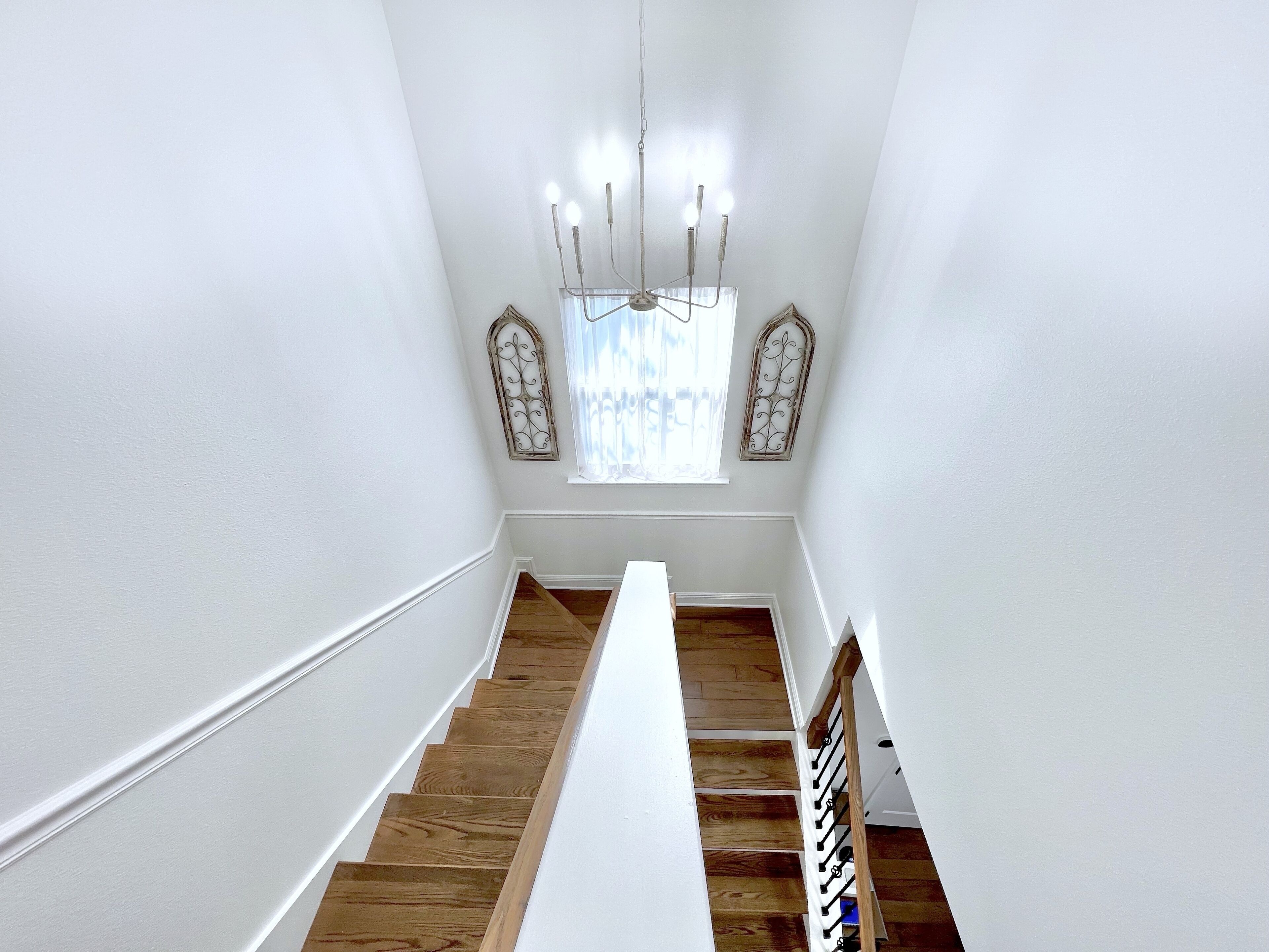 Una escalera de madera con barandilla blanca que lleva a un rellano con una araña y marcos de ventanas decorativos.