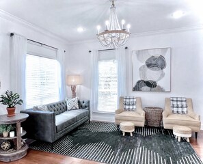 Un salon avec un canapé gris, deux fauteuils, un lustre, une grande peinture abstraite et un tapis à motifs.