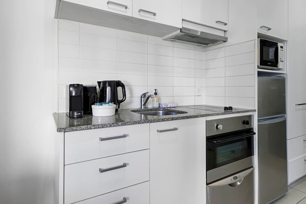 Una cocina moderna con armarios blancos, encimera de granito, cafetera, tetera, fregadero y horno y microondas integrados.