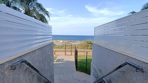 Ein Weg führt zu einem Strand mit Blick auf das Meer, gesäumt von weißen Wänden und Metallgeländern.