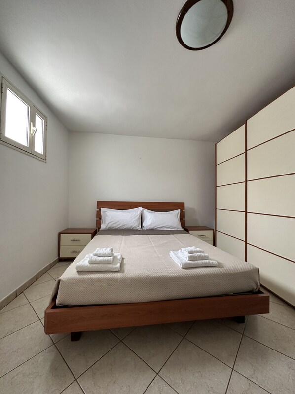 Una cama bien hecha con estructura de madera y ropa blanca, una mesita de noche con cajón, un armario con espejo y una ventana con persianas.