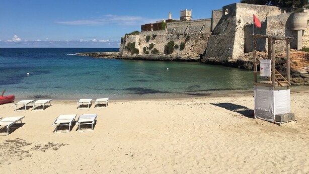 A beach with white lounge chairs, a stone wall, and a flag. - VILLA Indipendente con Piscina privata e accesso al mare. (Puglia)