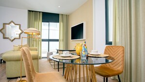Quarto de hotel moderno com uma mesa de vidro redonda, duas cadeiras, um sofá, um abajur e uma TV fixada na parede.